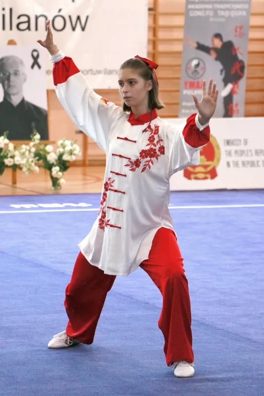 Kobieta w biało-czerwonym stroju stoi z rozstawionymi rękami i nogami. Prezentuje prawdopodobnie przejście między jedną pozycją, a drugą.